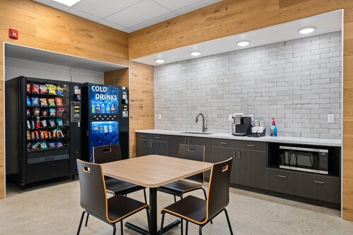 The image depicts a modern break room or kitchenette area, likely found in an office or workplace setting. The space features a clean and contemporary design with light wood paneling on the walls and a white tiled backsplash. The lighting is bright and evenly distributed, creating a welcoming atmosphere.

Prominently, there is a vending machine on the left side, stocked with various snacks and a separate section for cold drinks, indicated by the "COLD DRINKS" label. Next to the vending machine is a countertop area with dark cabinetry. On the countertop, there is a coffee maker and a bottle of dish soap, suggesting the area is equipped for basic food and beverage preparation.

In the foreground, there is a small square table with four chairs, designed with a minimalist aesthetic. The chairs have a dark finish with light edges, complementing the overall color scheme of the room.

The setting conveys a functional and inviting space designed for relaxation or casual meetings, typical of a break room environment. The use of neutral colors and modern fixtures suggests a professional and organized atmosphere.