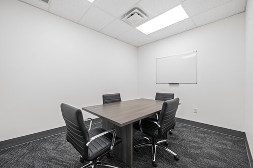 The image depicts a small, modern conference room with a minimalist design. The room features a rectangular wooden table surrounded by four black leather office chairs on wheels. The chairs have a sleek, contemporary design with horizontal stitching on the backrests. The room's walls are painted white, contributing to a clean and professional atmosphere.

On one wall, there is a whiteboard, suggesting the room is used for meetings, brainstorming sessions, or presentations. The floor is covered with a dark gray carpet, adding a touch of contrast to the otherwise light-colored space.

The ceiling is fitted with recessed lighting panels, providing bright, even illumination throughout the room. The overall atmosphere is one of professionalism and functionality, suitable for business meetings or small group discussions. The setting is typical of a corporate office environment, emphasizing efficiency and focus.
