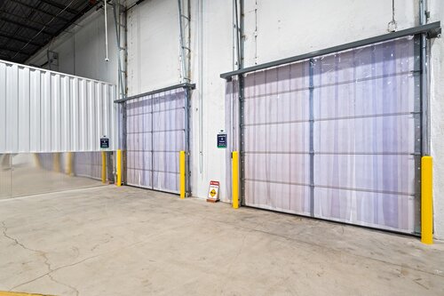 The image depicts an industrial setting, likely a warehouse or storage facility. It features two large, closed roll-up doors, each framed by yellow safety bollards. The doors are metallic with a semi-transparent appearance, suggesting they might be made of a lightweight material like plastic or thin metal. The floor is concrete, showing some wear and cracks, indicative of heavy use.

The walls are painted white, contributing to a clean and utilitarian atmosphere. Above the doors, there are visible tracks and mechanisms for opening and closing the doors. The ceiling is high, with exposed beams and industrial lighting, reinforcing the warehouse setting.

There are small signs on each door, possibly indicating instructions or safety information, but the text is not legible. A caution sign is placed on the floor between the doors, likely warning of potential hazards in the area.

The lighting is bright and even, suggesting overhead fluorescent lights typical in such environments. The overall atmosphere is functional and orderly, with an emphasis on practicality and safety. There are no people or other activities visible, focusing the image on the architectural and industrial elements.
