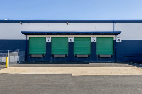 The image depicts a series of loading docks, numbered 4 through 7, on the exterior of an industrial or warehouse building. The building facade is painted in a combination of white and dark blue, creating a clean and orderly appearance. Each dock features a large green overhead door, indicating areas for loading and unloading goods. Above each door, the numbers are prominently displayed in black on white backgrounds, aiding in identification and organization.

The setting is likely a commercial or industrial area, with a focus on logistics or distribution. The ground in front of the docks is paved with asphalt, and there is a small concrete ramp leading up to the dock level, facilitating vehicle access. A chain-link fence with a yellow bollard is visible on the left side, suggesting security measures are in place.

The lighting is bright and even, suggesting the photo was taken during the day under clear skies. The overall atmosphere is utilitarian and functional, emphasizing efficiency and organization. The image conveys a sense of readiness and capability, typical of facilities involved in supply chain operations.