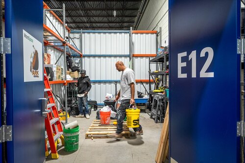 The image depicts an industrial storage or workshop area, likely within a warehouse or similar facility. The setting is framed by two blue doors, with the one on the right prominently labeled "E12" in white text. This suggests an organized system of storage or workspaces, typical in large facilities.

Inside the room, two individuals are engaged in work-related activities. One person, wearing a white shirt, is walking while carrying two buckets—one orange and one yellow—both labeled with brand names, indicating they might contain construction materials or tools. The other individual, dressed in darker clothing, is positioned towards the back, possibly organizing or handling equipment.

The room is well-lit, with overhead lighting casting a bright, functional atmosphere. The walls are lined with metal shelving units filled with various items, including a red ladder, toolboxes, and other industrial supplies. The floor is concrete, adding to the utilitarian feel of the space.

The overall atmosphere is one of productivity and organization, characteristic of a work environment focused on tasks such as maintenance, construction, or logistics. The presence of branded buckets and organized shelving suggests a professional setting where efficiency and order are prioritized.