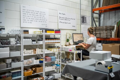 The image depicts a workspace within what appears to be a small warehouse or storage area, likely used for a small business or startup. The setting is organized and functional, with a focus on inventory and administrative tasks. 

A woman is seated at a desk, working on a computer. She is facing a wall, which has several whiteboards or posters with text on them. The text on the boards includes motivational quotes and business-related notes, suggesting a focus on productivity and inspiration. The woman is casually dressed, indicating a relaxed work environment.

Surrounding her are shelves filled with various products, including bottles, boxes, and jars, all neatly arranged. The products are likely part of the business inventory, possibly related to cosmetics, health, or wellness, given the appearance of the containers. There are also cardboard boxes and packaging materials on the shelves and a nearby table, indicating that this space is used for packing and shipping products.

The lighting is bright and utilitarian, typical of a workspace designed for efficiency. The color palette is neutral, with whites, grays, and natural tones dominating the scene, contributing to a clean and organized atmosphere.

Overall, the image conveys a sense of industriousness and entrepreneurship, capturing the essence of a small business operation focused on product fulfillment and office work.