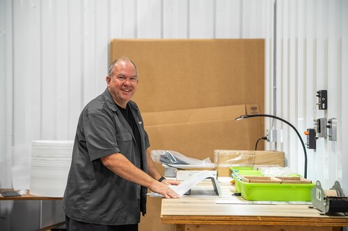 The image depicts a man standing in a workspace that appears to be a warehouse or workshop. He is smiling and engaged in an activity that involves handling materials on a wooden table. The man is wearing a dark gray shirt and is positioned near stacks of white sheets or boards, possibly indicating an industrial or manufacturing setting.

The table is organized with various items, including a green tray containing smaller objects or tools, and a desk lamp providing focused lighting on the workspace. The background features large cardboard sheets leaning against a white corrugated wall, suggesting a storage or packing area.

The lighting is bright and evenly distributed, contributing to a professional and industrious atmosphere. The overall color palette is neutral, with whites, grays, and natural wood tones dominating the scene.

The setting conveys a sense of productivity and organization, typical of environments where assembly, packing, or quality control might take place. The man's expression and posture suggest a positive and engaged attitude towards his work.