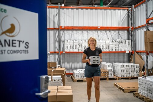 The image depicts a woman in a warehouse setting, carrying a stack of small boxes. She is wearing a black t-shirt and shorts, and she appears to be smiling, suggesting a positive or productive atmosphere. The warehouse is organized, with metal shelving units filled with more similar boxes in the background. The floor is concrete, typical of industrial or storage spaces.

On the left side of the image, a blue door is partially open, displaying a logo with a bird and the text "PLANET'S" which suggests the name of a company or brand, possibly related to composting or eco-friendly products, as inferred from the word "COMPOSTES" visible on the door. The lighting is bright, indicating a well-lit environment, possibly due to overhead industrial lights.

The setting conveys a sense of industriousness and organization, with a focus on logistics or product handling. The presence of the logo and text hints at an environmentally conscious business, aligning with themes of sustainability. The overall mood is one of efficiency and purpose, with the woman actively engaged in her task.