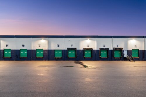 The image depicts a row of loading docks at a warehouse or industrial facility, captured during twilight. The setting features a series of large, green overhead doors, each numbered from 5 to 11, which are evenly spaced along the white and dark blue exterior wall of the building. Above each door, bright industrial lights illuminate the area, casting a warm glow that contrasts with the cool tones of the early evening sky. The sky transitions from a deep blue at the top to a lighter hue near the horizon, suggesting either dawn or dusk.

The ground in front of the loading docks is a wide, empty asphalt surface, slightly reflective, possibly due to recent rain or dew. The overall atmosphere is calm and quiet, typical of an industrial area outside of active working hours. There are no people or vehicles present, emphasizing a sense of stillness and solitude.

The setting and composition evoke themes of industrial efficiency and order, with the numbered doors suggesting organized logistics operations. The lighting and time of day contribute to a serene yet functional ambiance, characteristic of a well-maintained commercial facility. The image does not reference any specific cultural or historical context but is indicative of modern industrial architecture and operations.