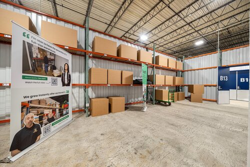 The image depicts the interior of a warehouse or storage facility. The setting features high ceilings with exposed metal beams and a concrete floor. Along the left side, there are metal shelving units filled with large cardboard boxes, some of which are sealed and others open. The shelves are organized, suggesting a systematic storage arrangement.

In the foreground on the left, there is a prominent vertical banner displaying photographs of individuals, likely employees, engaged in work-related activities. The banner includes text, although the specifics are not clear, it appears to emphasize teamwork or company values. The banner's design includes green accents, adding a touch of color to the otherwise industrial setting.

The background shows a door labeled "B13" and another labeled "B12," indicating different sections or storage areas within the facility. The lighting is bright and evenly distributed, creating a clean and organized atmosphere.

The overall mood of the image is professional and efficient, conveying a sense of order and productivity. The presence of the banner suggests a focus on corporate identity or employee recognition, common in business environments. The image captures the essence of a well-maintained storage or logistics operation, highlighting its organized and systematic nature.

[AI]
The image depicts an organized storage facility or warehouse with a focus on a section of shelving filled with large cardboard boxes. The shelves are made of metal and are arranged in a grid-like pattern, extending from the floor to the ceiling. The boxes are uniformly placed, suggesting a systematic approach to storage.

In the foreground, there is a prominent banner featuring photographs of individuals, possibly employees, along with text that includes a quotation and company branding. The banner appears to promote a business, emphasizing values such as growth and community. The individuals on the banner are wearing uniforms, which might indicate their association with the company being advertised.

The setting is well-lit with overhead industrial lighting, casting a bright and even illumination throughout the space. The walls and ceiling are constructed from corrugated metal, typical of industrial or storage environments.

On the right side of the image, there are two doors labeled "B13" and "B12," which might indicate different sections or storage units within the facility. The floor is made of concrete, adding to the industrial feel of the environment.

The overall atmosphere of the image is professional and organized, conveying a sense of efficiency and reliability. The presence of the banner suggests a marketing or promotional context, possibly for a logistics or storage company. The clean and orderly arrangement of the boxes and the clear labeling of sections contribute to the impression of a well-managed operation.