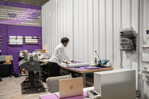 The image depicts an interior workspace with a person engaged in a task, possibly related to design or printing. The individual, dressed in a white shirt, is standing at a table covered with various materials, including rolled-up papers, suggesting an activity involving planning or production. The workspace features a predominantly purple and white color scheme, contributing to a professional and creative atmosphere.

On the left side, there is a large printer or plotter, indicating that this could be a print shop or a design studio. The walls are lined with shelves holding rolls of paper, reinforcing the theme of printing or graphic design. Above the workspace, there is a prominent purple wall with text, although the specific content of the text is not clearly visible.

A laptop and other office supplies are present on the tables, adding to the impression of a functional and busy work environment. The lighting is bright and even, enhancing visibility and focus on the tasks at hand. The overall setting conveys a sense of productivity and creativity, with a modern and organized aesthetic.

[AI]
The image depicts an interior workspace with a focus on a person engaged in a task. The setting appears to be a modern office or studio with a creative or technical focus. The individual, dressed in a white shirt, is standing at a desk, handling rolled-up paper, suggesting involvement in design, drafting, or planning activities.

The room is characterized by a clean, organized appearance with a prominent use of purple and white colors. The walls are white, with a section featuring a large purple panel that displays text. This text includes a quote about perseverance and vision, attributed to Henry David Thoreau, adding an inspirational or motivational theme to the environment.

The workspace is equipped with various office supplies and equipment. There is a printer or plotter, a laptop, and a desk organizer, indicating a professional setting that likely involves printing or graphic design work. The lighting is bright and even, contributing to a focused and productive atmosphere.

Overall, the image conveys a sense of diligence and creativity, with a modern and efficient workspace designed to inspire and facilitate detailed work.