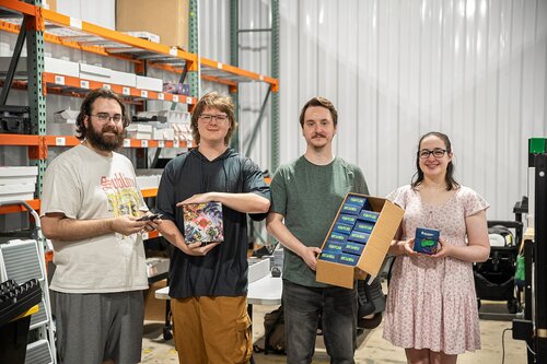 The image depicts four individuals standing in what appears to be a warehouse or storage area. The setting includes metal shelving stocked with various items, contributing to an industrial or commercial atmosphere. The walls are metallic, enhancing the utilitarian feel of the space.

The individuals are standing side by side, each holding different items related to card games. From left to right:

1. The first person is wearing a light-colored T-shirt with a graphic design and holding a pair of scissors.
2. The second person is wearing a dark shirt and holding a colorful box featuring fantasy artwork, possibly a card game or board game box.
3. The third person is wearing a green shirt and holding an open box displaying multiple packs of cards, suggesting they are part of a collectible card game.
4. The fourth person is wearing a light, patterned dress and holding a smaller box, which might also be related to card games.

The predominant colors in the image are muted, with the individuals' clothing providing some contrast against the neutral background. The lighting is bright and even, typical of indoor settings with overhead lighting.

The overall atmosphere is casual and friendly, suggesting a collaborative or hobbyist environment, possibly related to gaming or a small business operation involving card games. The image conveys a sense of community and shared interest in gaming or collectible card activities.

[AI]
The image depicts four individuals standing in what appears to be a warehouse or storage area. The setting includes metal shelving filled with various boxes and items, suggesting an environment related to storage, logistics, or retail. The walls are made of corrugated metal, contributing to an industrial atmosphere.

The individuals are positioned in a line, each holding different objects. From left to right:

1. The first person is wearing a light-colored T-shirt with text and graphics, holding a pair of scissors in one hand.
2. The second person is wearing a dark-colored shirt and is holding a colorful box with a fantasy-themed design, possibly a board game or a collectible item.
3. The third person is dressed in a green shirt and is holding an open box containing multiple smaller boxes, which could be card packs or similar items.
4. The fourth person is wearing a light-colored, patterned dress and is holding a tin or canister with a colorful design.

The predominant colors in the image are neutral tones from the background and clothing, with pops of color from the items being held. The lighting is bright and even, typical of indoor settings with overhead lighting.

The mood is casual and collaborative, suggesting a group activity or project related to games, collectibles, or retail products. The presence of game-related items hints at a possible connection to board games, trading card games, or similar hobbies. The individuals appear engaged and content, contributing to a positive and focused atmosphere.