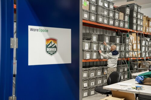 The image depicts an indoor storage or warehouse setting. On the left, a blue door is partially open, displaying a sign with the text "WareSpace" and a logo featuring a mountain design with the word "DENVER" and "OUTDOOR" underneath. This suggests a connection to outdoor activities or products, possibly in Denver.

Inside the warehouse, a man is organizing or retrieving items from stacked gray storage bins that are neatly arranged on metal shelves. The bins are labeled, indicating a systematic storage system. The man is wearing casual attire, including a hoodie and pants, and is focused on his task.

In the foreground, a white table is visible, cluttered with various items, including a green fabric, suggesting ongoing work or sorting activities. A black office chair is positioned nearby, indicating a workspace setup.

The lighting is bright and functional, typical of a workspace environment, creating a professional and organized atmosphere. The overall scene conveys a sense of efficiency and order, likely related to inventory management or preparation for outdoor-related activities or products.

[AI]
The image depicts an indoor warehouse setting with a focus on organization and storage. On the left side, there is a prominent blue door with a sign that reads "WareSpace" and features a logo with the word "DENVER" and a mountain design, suggesting a connection to Denver or a related organization.

In the background, a person is engaged in organizing or retrieving items from a series of stacked gray storage bins. These bins are neatly arranged on metal shelving units that reach the ceiling, indicating a well-structured storage system. The individual is wearing casual attire, including a hoodie and pants, and appears to be focused on their task.

In the foreground, there is a white table with various items on it, including papers and a green fabric, possibly used for packaging or covering. The setting suggests a functional and efficient workspace, likely used for inventory management or distribution.

The lighting is bright and even, contributing to a clean and orderly atmosphere. The overall theme of the image conveys efficiency, organization, and a professional environment, possibly within a logistics or storage facility. The presence of the Denver logo may indicate a regional focus or branding associated with the location or company.