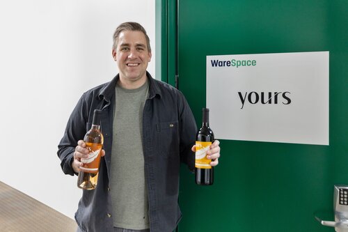 The image features a man standing in front of a green door with a sign that reads "WareSpace yours." He is smiling and holding two bottles of wine, one in each hand. The bottle on the left appears to be a rosé, while the one on the right is likely a red wine. The man is dressed casually in a gray jacket over a dark shirt and jeans. The setting suggests a modern, possibly industrial or office environment, with clean lines and a minimalist aesthetic. The lighting is bright and even, contributing to a cheerful and welcoming atmosphere. The presence of the "WareSpace" branding might indicate a connection to a business or service related to storage or logistics. The image conveys a sense of casual professionalism and hospitality.