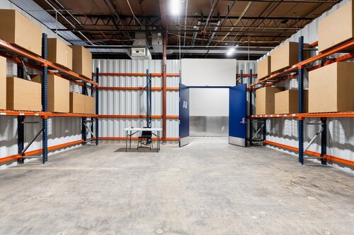 The image depicts a spacious storage room or warehouse with a utilitarian and industrial atmosphere. The room is lined with tall metal shelving units on both sides, painted in a bright orange color, which adds a pop of vibrancy to the otherwise neutral setting. These shelves are stocked with large, uniform cardboard boxes, suggesting an organized storage system.

The floor is concrete, typical of industrial spaces, and the walls are made of corrugated metal, reinforcing the warehouse aesthetic. Overhead, the ceiling is exposed, revealing a network of pipes and electrical conduits, contributing to the industrial feel.

At the center of the room, there is a small table with a chair, indicating a possible workspace or a spot for quick tasks. The table is simple and functional, aligning with the utilitarian theme of the room.

At the far end of the room, there are double doors painted in a bold blue color, which stand out against the neutral tones of the walls and floor. The doors are slightly ajar, suggesting a sense of openness or transition to another area.

The lighting is bright and evenly distributed, likely from overhead fluorescent lights, creating a well-lit environment conducive to work and storage activities. The overall atmosphere is one of efficiency and organization, typical of a space designed for logistical operations or inventory management.