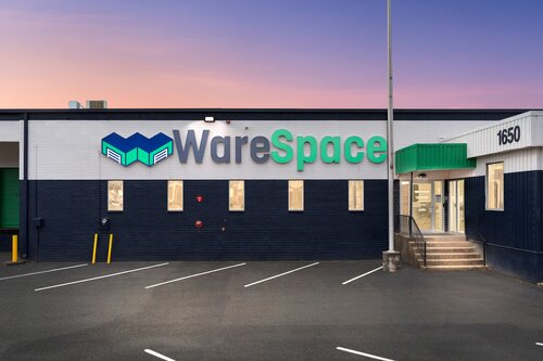 The image depicts the exterior of a commercial building, likely a warehouse or storage facility, named "WareSpace." The building's facade is painted in a combination of dark blue and white, with the name "WareSpace" prominently displayed in large, modern lettering. The word "Ware" is in blue, while "Space" is in green, and the logo features a stylized design that resembles a warehouse structure, incorporating the same blue and green colors.

The setting appears to be during twilight or early evening, as indicated by the soft, warm hues of the sky transitioning from pink to purple. The lighting is subtle, with exterior lights illuminating the entrance area. The building has a simple, industrial design with several rectangular windows and a visible entrance with steps leading up to it, marked by a green awning.

The number "1650" is clearly visible on the building, likely indicating the street address. The parking area in front of the building is empty, with clearly marked parking lines and a couple of yellow bollards near a garage door.

The overall atmosphere is professional and orderly, suggesting a business environment focused on storage or logistics. The clean and modern aesthetic, combined with the distinct branding, conveys a sense of reliability and efficiency.