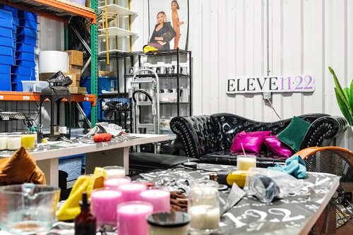 The image depicts an indoor setting that appears to be a workshop or creative studio, characterized by a mix of industrial and casual elements. The environment is cluttered with various objects and materials, suggesting ongoing projects or activities. 

Prominent in the scene are two tables covered with a variety of items, including candles of different colors and sizes, such as pink and white, and various crafting supplies. There are also tools and containers scattered across the surfaces, indicating a focus on candle-making or a similar craft. 

A black, tufted leather couch is positioned against a corrugated metal wall, adding a touch of comfort amidst the industrial surroundings. Brightly colored pillows, including pink and green, are placed on the couch, providing a vibrant contrast to the otherwise utilitarian setting.

In the background, shelves filled with blue storage bins and other materials suggest organization amidst the creative chaos. A ladder and a metal cart with additional supplies are also visible, indicating a space that is actively used for production or artistic endeavors.

On the wall, a sign reads "ELEVE11:22," which could be the name of the studio, a brand, or a thematic element related to the activities taking place. Nearby, a cutout of a person stands, adding a personal or promotional touch to the environment.

The lighting is bright and functional, typical of a workspace, and the overall atmosphere conveys a sense of industrious creativity and casual productivity. The setting suggests a place where art, craft, or small-scale manufacturing takes place, possibly for a niche market or personal business.