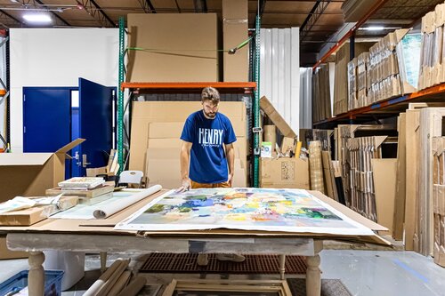 The image depicts a man standing in what appears to be an art studio or workshop. He is focused on a large, colorful abstract painting laid out on a table in front of him. The painting features a mix of vibrant colors, including yellows, blues, greens, and reds, creating a dynamic and lively composition.

The man is wearing a blue T-shirt with the word "HENRY" printed in bold white letters. He appears to be examining or working on the painting, suggesting an artistic or creative process. The setting is an industrial space with high ceilings, exposed beams, and shelves filled with various materials and supplies, including cardboard, packaging materials, and other artworks or canvases.

The lighting is bright and evenly distributed, highlighting the workspace and the painting. The atmosphere conveys a sense of creativity and industriousness, typical of an artist's studio where creation and preparation are ongoing.

The environment suggests a focus on art production or preparation, possibly for an exhibition or sale. The presence of packaging materials and the organized yet cluttered space indicate an active workspace where art is both created and prepared for distribution.
