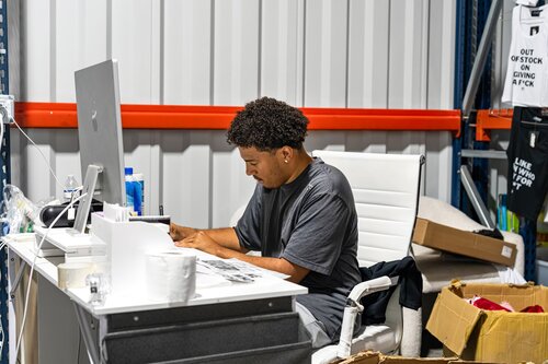 The image depicts a young man seated at a white desk in what appears to be a warehouse or industrial setting. He is focused on his work, looking down at papers or a screen in front of him. The individual is wearing a dark gray t-shirt and has curly hair. The desk is equipped with a large computer monitor, various papers, and office supplies, suggesting a workspace environment.

Behind the desk, there is a corrugated metal wall, indicating an industrial or storage facility. A red horizontal beam runs across the wall, adding a splash of color to the otherwise neutral setting. To the right, there are cardboard boxes, some open, which might imply inventory or shipping activities.

On the wall, there is a black shirt with white text that reads, "OUT OF STOCK ON GIVING A F**K," adding a humorous or irreverent touch to the atmosphere. This text suggests a casual or laid-back work environment. The lighting is bright, likely from overhead industrial lights, creating a well-lit workspace.

The overall atmosphere is one of focused productivity, with a hint of humor and informality suggested by the text on the shirt. The setting and elements indicate a modern, possibly startup or small business environment, where casual dress and humor are part of the workplace culture.
