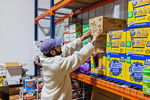 The image depicts a person in a storage or warehouse setting, reaching up to organize or retrieve items from a shelf. The individual is wearing a light-colored fleece jacket and a purple cap, suggesting a casual or work-related environment. The setting is brightly lit, with industrial shelving units filled with various packaged goods.

Prominent in the scene are multiple boxes of "Linden's Butter Crunch" cookies, which are vividly colored in blue and yellow packaging. These boxes are stacked neatly on the shelves, indicating a focus on inventory or stock management. Other visible items include boxes of "Grandma's Cookies" and other assorted snacks, adding to the impression of a food storage area or distribution center.

The overall atmosphere is functional and industrious, with a sense of organization and efficiency. The setting suggests a backroom of a grocery store, a warehouse, or a similar environment where goods are stored and managed. The presence of branded snack products might indicate a commercial or retail context.

The image conveys a routine activity, likely related to stocking or inventory management, and the colorful packaging adds a lively visual element to the otherwise utilitarian space.
