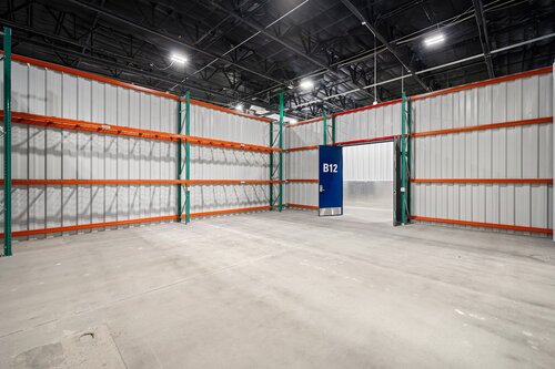 The image depicts an empty industrial warehouse space with a focus on its structural elements. The setting features high ceilings with exposed beams and a grid of overhead lighting, casting a bright, even illumination across the area. The walls are constructed of corrugated metal panels, and the floor is a smooth, polished concrete, suggesting a modern and utilitarian design.

Prominent in the image are the metal shelving units lining the walls, painted in a combination of green and orange, which are common colors for industrial shelving due to their visibility and durability. These shelves are currently empty, emphasizing the spaciousness and potential for storage or other industrial activities.

A notable feature is a blue door labeled "B12," which stands out against the otherwise neutral color palette. This door may indicate a specific section or storage area within the warehouse, suggesting organization and compartmentalization typical of large storage facilities.

The overall atmosphere of the image is one of cleanliness, order, and readiness for future use, typical of a newly constructed or recently renovated industrial space. The setting does not convey any specific cultural or historical context but is representative of contemporary warehouse design, often seen in logistics, manufacturing, or large-scale retail operations.

[AI]
The image depicts a large, empty warehouse space with a focus on industrial shelving and a prominent blue door labeled "B12." The warehouse features high ceilings with exposed beams and lighting fixtures, casting a bright, even light across the space. The walls are made of corrugated metal panels, contributing to the industrial aesthetic.

The shelving units are constructed with green metal frames and orange horizontal beams, suggesting they are designed for heavy-duty storage. The absence of items on the shelves emphasizes the spaciousness and potential for storage or industrial use.

The floor is made of smooth concrete, typical for warehouses, and the overall color palette is neutral, dominated by grays and metallic tones, with the blue door providing a contrasting focal point.

The atmosphere is utilitarian and orderly, conveying a sense of readiness for future use, whether for storage, manufacturing, or distribution. The setting suggests a modern, well-maintained facility, possibly newly constructed or recently renovated, given its pristine condition.

There are no visible people or activities taking place, reinforcing the impression of an unoccupied or newly prepared space. The image does not reference any specific cultural, historical, or entertainment context, focusing instead on the functional and architectural elements of the warehouse environment.