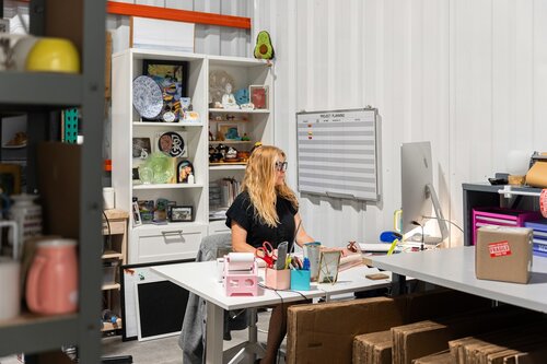 The image depicts a woman seated at a desk in a workspace that appears to be a combination of an office and a storage area. She is working on a computer, with her attention focused on the screen. The setting is well-lit, with a mix of natural and artificial lighting, creating a bright and organized atmosphere.

The woman has long blonde hair and is wearing glasses and a black shirt. The desk is cluttered with various office supplies, including a pink tape dispenser, a pen holder, and a stack of papers. A computer monitor is prominently placed on the desk, and a cardboard box with a "Thank You" sticker is visible, suggesting a business or shipping-related activity.

Behind her, there is a white shelving unit filled with various items, such as decorative objects, books, and a large plate. A whiteboard is mounted on the wall, displaying a schedule or list, indicating a structured work environment.

The predominant colors in the image are neutral tones, with pops of color from the objects on the shelves and desk. The overall atmosphere is one of productivity and organization, typical of a small business or creative workspace. The presence of packing materials and boxes suggests an e-commerce or shipping-related business.

[AI]
The image depicts a workspace within what appears to be a storage or warehouse setting. A woman with long blonde hair is seated at a desk, working on a computer. She is wearing a black shirt and glasses, and her attention is focused on the screen. The desk is cluttered with various office supplies, including a pink tape dispenser, a pen holder, and a box labeled "Priority Mail."

Behind her, there is a white shelving unit filled with an eclectic mix of items, such as decorative plates, figurines, and a small globe. A notable feature is a large avocado-shaped object placed on top of the shelf. A whiteboard with a "Weekly Planner" is mounted on the wall to her right, containing a grid layout but no visible text.

The environment is brightly lit, suggesting a productive atmosphere. The walls are made of corrugated metal, typical of industrial or storage spaces. In the foreground, there are cardboard boxes stacked on the floor, indicating that this might be a packing or shipping area.

The overall scene conveys a sense of industriousness and organization amidst a casual and personalized workspace. The presence of various decorative items adds a touch of individuality and creativity to the otherwise utilitarian setting.