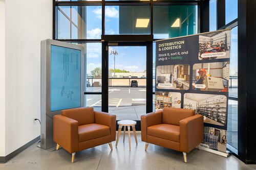The image depicts a modern, professional waiting area or lobby, characterized by a clean and organized setting. Two contemporary, brown leather armchairs are positioned symmetrically around a small, round white table, creating a welcoming and comfortable seating arrangement. The area is well-lit, with natural light streaming in through large floor-to-ceiling windows, which also offer a view of the outside parking lot and a clear blue sky.

To the left, there is a large digital display or interactive screen, possibly used for informational or advertising purposes. On the right, a vertical banner stands prominently, featuring the text "DISTRIBUTION & LOGISTICS" along with the tagline "Stock it, sort it, and ship it – reliably." The banner includes several images related to logistics, such as warehouse operations, shelving, and machinery, conveying a theme of efficiency and modernity in logistics services.

The overall atmosphere is professional and business-oriented, suggesting a setting related to corporate offices, a logistics company, or a similar business environment. The use of neutral colors like brown and white, combined with the bright natural lighting, creates a calm and inviting ambiance. The design elements and decor suggest a focus on functionality and modern aesthetics, typical of contemporary business spaces.

[AI]
The image depicts a modern, professional waiting area or lobby, likely within a business or office setting. Two brown leather armchairs are positioned facing each other with a small, round white table between them. The armchairs have a sleek, contemporary design, contributing to a clean and inviting atmosphere.

Behind the chairs, a large window provides natural light, enhancing the open and airy feel of the space. The view outside shows a parking lot and some greenery, suggesting the building is located in a suburban or commercial area.

To the left, there's a tall, rectangular digital display or kiosk, which appears to be inactive or turned off, as the screen is a solid blue. On the right, a large vertical banner stands prominently. The banner features images and text related to "Distribution & Logistics," with phrases like "Stock it, sort it, and ship it – flexibly." The images on the banner depict various aspects of logistics, such as a warehouse setting, a worker operating machinery, and shelving with products.

The color palette is dominated by neutral tones, with the brown of the chairs and the white of the table complemented by the black and gray elements of the window frames and flooring. The lighting is bright and natural, contributing to a professional and welcoming environment.

Overall, the setting conveys a sense of efficiency and modernity, likely intended to reflect the business's focus on logistics and distribution. The atmosphere is calm and organized, suitable for a corporate or industrial context.