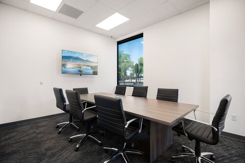 The image depicts a modern conference room with a minimalist design. The room features a long rectangular wooden table surrounded by eight black leather office chairs on wheels. The chairs have a sleek, contemporary design with horizontal stitching. 

On the wall to the left, there is a landscape painting showcasing a serene scene with mountains and a body of water, adding a touch of color and tranquility to the otherwise neutral space. The predominant colors in the room are shades of black, white, and gray, creating a professional and clean atmosphere.

A large window on the right side of the room allows natural light to filter in, offering a view of trees and a clear sky outside, which enhances the room's openness and brightness. The carpet is dark gray, complementing the overall color scheme.

The ceiling has recessed lighting, providing ample illumination, and the walls are painted white, contributing to the room's airy and spacious feel. The setting suggests a corporate or business environment, suitable for meetings, discussions, or presentations. The overall atmosphere is calm, professional, and conducive to focused work or collaboration.

[AI]
The image depicts a modern conference room with a minimalist design. The room features a long rectangular wooden table surrounded by eight black leather office chairs on wheels. The chairs have a sleek, contemporary design with horizontal stitching on the backrests. The floor is covered with dark gray carpet, adding a professional and subdued tone to the space.

On one wall, there is a large window that allows natural light to flood the room, providing a view of greenery outside, which adds a touch of nature to the otherwise corporate setting. The opposite wall displays a landscape painting, featuring a serene scene with mountains and a body of water, contributing to a calming atmosphere.

The walls and ceiling are painted white, enhancing the brightness and spaciousness of the room. The ceiling has recessed lighting panels, ensuring the room is well-lit even without natural light. The overall ambiance is professional and tranquil, suitable for meetings or collaborative work sessions.

The setting suggests a corporate environment, likely within an office building, designed for functionality and comfort. The combination of natural elements with modern furnishings creates a balanced and inviting atmosphere.