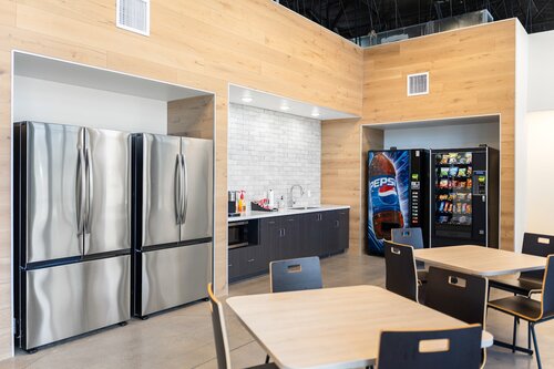 The image depicts a modern break room or kitchenette area, likely within an office or corporate setting. The room features a clean and organized design with light wood paneling on the walls, creating a warm and inviting atmosphere. 

Prominent in the image are two large stainless steel refrigerators, positioned side by side on the left. These appliances contribute to a sleek and contemporary aesthetic. Adjacent to the refrigerators is a small kitchenette area with a white subway tile backsplash, a countertop, and a sink, suggesting functionality for meal preparation or cleanup.

On the right side of the image, there is a vending machine with a prominent Pepsi logo, offering various snacks and beverages. Next to it is another vending machine, likely for additional snacks or drinks, indicating convenience for employees or visitors.

In the foreground, several tables and chairs are arranged, suggesting a space for dining or casual meetings. The furniture is simple and functional, with light-colored tabletops and dark chairs, maintaining the room's modern theme.

The lighting is bright and evenly distributed, enhancing the room's clean and professional look. The overall atmosphere is one of efficiency and comfort, designed to accommodate breaks and informal gatherings in a workplace environment.

[AI]
The image depicts a modern break room or kitchen area, likely in an office or workplace setting. The room features a clean and contemporary design with light wood paneling on the walls and a neutral color palette. 

On the left side, there are two large stainless steel refrigerators, suggesting ample storage space for food and beverages. In the center, there is a kitchenette area with a white brick backsplash, a countertop, and a sink. Various items, such as a coffee maker and other small appliances, are placed on the counter, indicating the space is equipped for meal preparation or coffee breaks.

To the right, a vending machine with a prominent Pepsi logo is visible, offering snacks and drinks. Next to it, there is another vending machine, likely for additional snacks or beverages. 

In the foreground, several tables and chairs are arranged, providing seating for employees or visitors to sit and enjoy their meals or breaks. The furniture is simple and functional, matching the overall minimalist aesthetic of the room.

The lighting is bright and even, contributing to a welcoming and comfortable atmosphere. The setting suggests a focus on convenience and relaxation, typical of a communal area designed for employees to unwind during their workday.