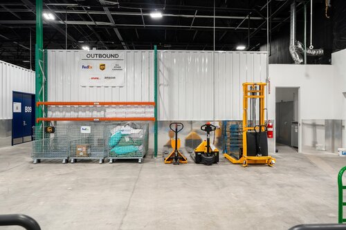 The image depicts an industrial or warehouse setting with a focus on storage and logistics. The environment is clean and organized, with a concrete floor and high ceiling, typical of a warehouse. 

Prominent in the image is a wall with a sign that reads "CUTESOUND," accompanied by logos of well-known courier companies, FedEx and UPS, suggesting a shipping or logistics operation. Below the sign, there are two large metal mesh storage containers filled with various materials, possibly packaging supplies or goods ready for shipment.

In front of the wall, there are two pallet jacks, one orange and one yellow, used for moving pallets within the warehouse. Next to them is a taller, yellow manual stacker, indicating the handling of heavier or bulkier items.

The lighting is bright and even, typical of industrial settings, providing a clear view of the space. The overall atmosphere is functional and efficient, emphasizing order and readiness for logistical operations.

The setting suggests a modern warehouse facility, possibly part of a distribution center or a fulfillment operation, where goods are processed and shipped. The presence of major courier logos indicates a focus on delivery and transportation services.

[AI]
The image depicts an industrial or warehouse setting with a focus on logistics and storage. The environment is clean and organized, featuring a concrete floor and high ceiling with visible black structural elements. The lighting is bright, likely from overhead fluorescent lights, contributing to a well-lit atmosphere.

Prominently displayed on the wall is a sign with the text "CUTESOUND" and logos of well-known shipping companies: FedEx, UPS, and DHL, suggesting a logistics or shipping hub. Below the sign, there are two large metal mesh storage bins. One of the bins contains teal-colored bags or packages, indicating organized storage or sorting of items.

In front of the bins, there are three pieces of equipment: two manual pallet jacks, one black and one yellow, and a yellow hydraulic stacker, which is used for lifting and moving pallets. These tools are essential for handling goods within a warehouse, highlighting the functional aspect of the space.

The walls are a combination of white and metallic surfaces, contributing to a modern industrial aesthetic. A partially open door on the right side leads to another area, possibly an office or storage room, suggesting the space is part of a larger facility.

Overall, the image conveys a sense of efficiency and order, typical of a professional logistics or distribution center. The presence of major shipping company logos emphasizes the global or large-scale nature of the operations conducted in this space.