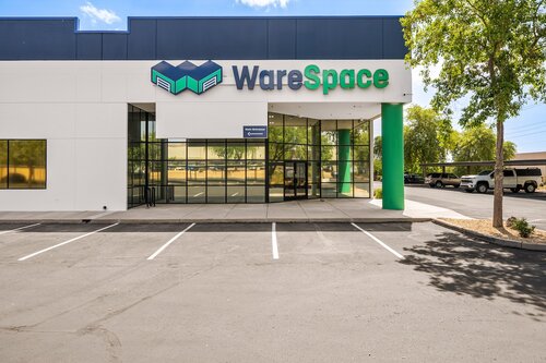 The image depicts the exterior of a modern commercial building, prominently featuring the name "WareSpace" in bold letters on the facade. The building has a sleek, contemporary design with a combination of white and dark blue colors. The logo, positioned above the text, consists of geometric shapes that resemble interconnected cubes, suggesting themes of storage or logistics.

The entrance is glass-fronted, allowing a view into the interior, which appears spacious and well-lit. A small sign near the entrance reads "Warehouse," indicating the building's function. The setting includes a well-maintained parking area with clearly marked spaces, and a few trees provide a touch of greenery, enhancing the urban environment.

The atmosphere is professional and business-oriented, likely associated with warehousing or storage services. The presence of clear skies and bright lighting suggests a daytime setting, contributing to a clean and inviting appearance. The overall scene conveys efficiency and modernity, typical of commercial spaces designed for business operations.

[AI]
The image depicts the exterior of a modern commercial building with a focus on a business named "WareSpace." The building features a sleek, contemporary design with a combination of white and dark blue colors. The "WareSpace" logo is prominently displayed at the top of the building, featuring a stylized geometric design in blue and green, suggesting a theme related to storage or logistics.

The entrance is made of glass, providing a clear view into the interior, which appears to be well-lit and spacious. The facade includes large windows that reflect the surrounding environment, including a clear blue sky and nearby trees, contributing to a bright and inviting atmosphere.

The foreground shows a neatly maintained parking area with clearly marked spaces, indicating a professional and organized setting. The presence of green pillars and surrounding trees adds a touch of nature to the otherwise urban environment.

Overall, the image conveys a sense of professionalism and modernity, likely associated with a business offering storage or workspace solutions. The setting suggests a clean, efficient, and customer-friendly environment.