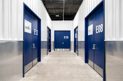 The image depicts a corridor in a storage facility, characterized by its industrial and utilitarian design. The setting features a series of storage unit doors, each painted in a striking blue color, with prominent white alphanumeric labels: E12, E10, and E08. These labels are clearly visible and likely indicate the unit numbers for organizational purposes.

The corridor is lined with metallic, diamond-patterned paneling along the lower half of the walls, contributing to the industrial aesthetic. The upper walls are white, enhancing the brightness and cleanliness of the space. The ceiling is black, with visible ducts and lighting fixtures, suggesting a warehouse-like environment.

The floor is made of polished concrete, adding to the functional and minimalist design. The lighting is bright and evenly distributed, creating a well-lit atmosphere that is typical of storage facilities, ensuring visibility and safety.

The overall atmosphere is orderly and efficient, designed for practicality rather than comfort. This setting is commonly associated with self-storage facilities where individuals or businesses rent space to store personal or commercial items. The image conveys a sense of organization and security, typical of environments where personal belongings are kept.

[AI]
The image depicts a hallway in a storage facility. The setting features a series of blue metal doors, each labeled with white alphanumeric identifiers such as "E12," "E10," and "E08." These identifiers suggest a systematic organization typical of storage units. The doors are evenly spaced along the white corrugated metal walls, which contribute to an industrial aesthetic.

The floor is concrete, enhancing the utilitarian feel of the environment. The lower portion of the walls is covered with a diamond plate metal, adding a textured, durable appearance. The ceiling is black, with visible industrial lighting fixtures, providing functional illumination to the space.

The overall atmosphere is orderly and efficient, characteristic of a storage or warehouse facility. The predominant colors are blue, white, and metallic silver, which create a clean and professional look. The setting is devoid of people, emphasizing the focus on the storage units themselves.

This image is likely to be used in contexts related to storage solutions, organization, or industrial facilities. The clear labeling and structured layout suggest a well-maintained and secure environment for storing personal or business items.