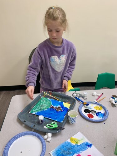 A young child is engaged in a painting activity at a table. The child, with light-colored hair tied back, is wearing a purple sweatshirt featuring a large, sequined heart design on the front. The setting appears to be an indoor classroom or art room with neutral-colored walls and a wooden floor. 

On the table, there is a variety of art supplies, including a paint palette with black, red, yellow, and white paint, a pair of green scissors, cotton balls, and a cup with a brush. The child is focused on a tray holding a painting that depicts a landscape with blue, green, and yellow colors. The mood is creative and educational, with a focus on art and expression. The lighting is bright and even, suggesting a well-lit environment conducive to detailed work. 

The scene captures a moment of concentration and creativity, emphasizing the child's involvement in the artistic process.
