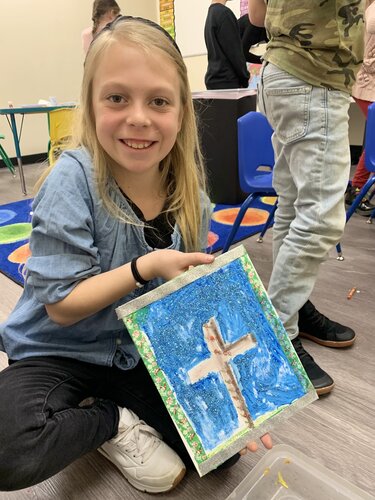 Young girl sitting on the floor in a classroom setting, holding a piece of artwork. The artwork features a cross in the center, surrounded by a blue background and a decorative border. She is wearing a light blue denim jacket over a black shirt, with black pants and white sneakers. Her hair is long and blonde. 

In the background, there are other children standing and engaging in activities, one wearing a camouflage t-shirt and jeans. The classroom has colorful circular rugs and blue chairs. The floor is wooden, and the walls are light-colored. The lighting is bright, creating a cheerful and creative atmosphere. The scene suggests an art or craft activity in an educational environment.