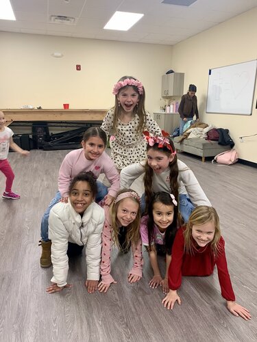 Group of seven children forming a human pyramid indoors. The setting is a room with light-colored walls and a wooden floor. The lighting is bright and even, creating a cheerful mood. 

The children are dressed in casual, colorful clothing. The top child wears a floral dress and a pink headband, smiling widely. The base of the pyramid consists of four children, two wearing light-colored tops and two in darker hues. One child in the middle wears a white jacket, while another sports a red sweater. 

In the background, a whiteboard is mounted on the wall, and there are various items like a backpack and coats on a bench. A person is partially visible, standing near the wall. The environment suggests a casual, playful gathering, possibly in a community or recreational center.