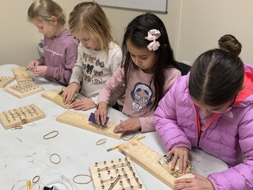 Group of four children engaged in a hands-on activity at a table. Each child is focused on assembling small wooden pegboards with rubber bands and nails. The children are dressed in casual clothing: one wears a pink jacket, another a lavender hoodie with visible text "Champion," a third in a light pink sweater with animal prints, and the fourth in a white shirt with a graphic design. The setting is a simple, neutral-colored room with a whiteboard partially visible in the background. The table is covered with various materials, including pencils, rubber bands, and additional pegboards. The lighting is bright and even, creating a focused and educational atmosphere. The mood is concentrated and collaborative, with the children appearing to be learning or participating in a workshop or class.