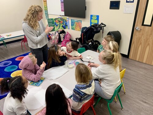 Classroom setting with a group of young children seated around a rectangular table. The children are engaged in an activity, focusing on a woman standing at the head of the table. She is holding a clear plastic bag containing colorful objects, possibly for a demonstration or lesson. The woman is wearing a gray sweater and jeans, with curly blonde hair.

The children, dressed in casual clothing, are seated on colorful chairs—red, yellow, and green. Some children have notebooks or papers in front of them, suggesting a learning activity. The classroom has a vibrant and playful atmosphere, with a colorful circular rug featuring multicolored circles visible in the background.

The walls are adorned with educational posters, including a map and other instructional materials. A stroller is parked against the wall, indicating a family-friendly environment. The lighting is bright and even, contributing to a cheerful and engaging mood. The room's layout is organized to facilitate interaction and learning.