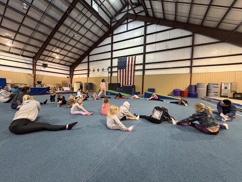 Indoor gymnasium setting with a high, metal-beamed ceiling and large American flag displayed on the wall. The scene features a group of children and adults engaged in a stretching exercise on a blue padded floor. The participants are dressed in casual athletic wear, including sweatshirts, leggings, and t-shirts, with some wearing socks. 

The group is spread out across the floor, each person performing various stretching poses, such as seated forward bends and leg stretches. The environment is well-lit with overhead lighting, creating a bright and focused atmosphere. In the background, gymnastic equipment like mats and stacked foam blocks are visible, suggesting a multi-purpose sports facility. The mood is active and focused, emphasizing physical activity and group participation.