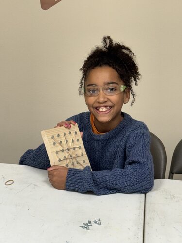 A young person is seated at a white table, smiling and holding a wooden board with a geometric pattern made of nails and string. They are wearing a blue knit sweater and clear safety glasses. The background is a plain, light-colored wall, creating a neutral setting. The table has a few scattered small metal objects, possibly nails or screws. The lighting is bright and even, suggesting an indoor environment. The mood is cheerful and engaging, with a focus on a creative or educational activity involving crafting or construction. The individual appears to be involved in a hands-on project, likely related to art or engineering.