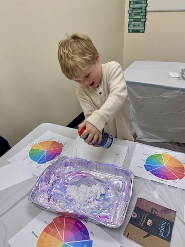 A young child with light hair is engaged in a hands-on activity. The child is wearing a light-colored, long-sleeved shirt. They are enthusiastically spraying a can of shaving cream into a rectangular aluminum tray placed on a table. The table is covered with a white cloth and is surrounded by several printed color wheels, which are visible on sheets of paper. The setting appears to be an indoor educational or creative environment, possibly a classroom or activity room, with neutral-colored walls. The lighting is bright and even, creating a cheerful and focused mood. A book with a visible cover featuring a shark is placed on the table, adding an element of interest. The overall scene suggests a playful and educational activity involving color exploration or sensory play.