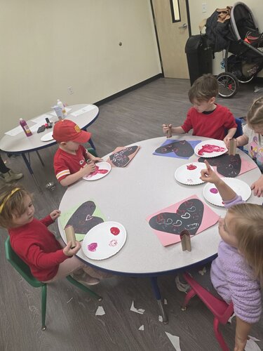 Children are seated around two small, white, kidney-shaped tables in a classroom setting, engaging in an art activity. The children, wearing casual clothing in shades of red and pink, are focused on creating heart-shaped crafts. Some are using paintbrushes to apply red and pink paint onto black heart-shaped paper. The tables are scattered with art supplies, including paint bottles, brushes, and paper plates used as palettes. The room has a neutral-colored wall and a closed wooden door in the background. A stroller is visible in the corner, suggesting a childcare environment. The lighting is bright and even, creating a cheerful and creative atmosphere. The floor is covered with gray carpeting, and small pieces of paper are scattered around, indicating active participation in the craft activity.