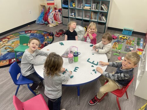 A group of young children are seated around a circular white table in a classroom setting. The children are engaged in a creative activity using green modeling clay, forming various shapes and letters. The visible text on the table includes the letters "E" and "M" made from the clay. The children are dressed in casual clothing, including sweaters and jeans. 

The classroom environment features a colorful, circular rug with a world map design in the background, along with shelves filled with books and toys. Additional toys, such as cars and a toy road mat, are scattered around the room. The walls are a neutral color, and the flooring is a light gray. 

The lighting is bright and evenly distributed, creating a cheerful and lively mood. The scene captures a playful and educational atmosphere, emphasizing creativity and learning.