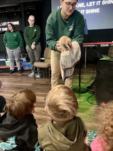People are gathered in an indoor setting with a focus on an educational or interactive demonstration. A person in a green sweatshirt and khaki pants is holding a small animal wrapped in a cloth, engaging with a group of children seated on the floor. The children are observing attentively, dressed in casual clothing, with some wearing hoodies. In the background, another person in a similar green sweatshirt stands with hands clasped, possibly assisting or observing the demonstration.

The environment features a wooden floor and a stage area with a keyboard visible, suggesting a multi-purpose room. On the wall, there is a projection screen displaying the text "LET IT SHINE, LET IT SHINE, LET IT SHINE," indicating a possible musical or thematic element to the event. The lighting is warm and inviting, creating an educational and interactive atmosphere. The mood is engaging and focused on learning and interaction with the animal.