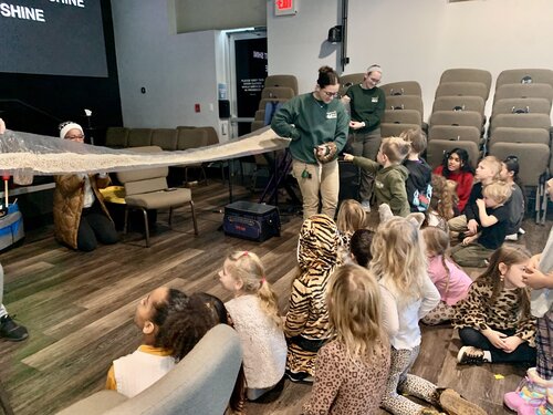 Audience of young children seated on a wooden floor in a semi-circle, facing a group of adults. The children are dressed in various outfits, including animal print clothing. The adults, wearing uniforms with name tags, appear to be educators or facilitators. They are holding a large, light-colored fabric or tarp, engaging the children in an interactive activity. 

The setting is an indoor room with rows of beige auditorium-style seats. The lighting is bright and even, creating a lively and educational atmosphere. On the wall, a large screen displays partial text: "SHINE" is prominently visible. The mood is educational and engaging, with the children appearing attentive and curious. 

Distinctive details include the presence of educational materials and equipment, such as a toolbox on the floor, suggesting a hands-on learning environment. The room's design and setup indicate a space used for presentations or workshops.