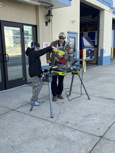 People: Two individuals are present. One is an adult wearing a tan jacket, blue jeans, and a tan cap. The other is a child wearing a black jacket, camouflage pants, and a black cap. The adult appears to be instructing or assisting the child.

Actions: The adult is guiding the child in operating a miter saw placed on a stand. The child is actively engaged, with hands on the saw.

Clothing: The adult is dressed in casual outdoor attire, including a tan jacket and cap. The child is wearing a black jacket, camouflage pants, and a black cap.

Objects: A yellow and gray miter saw is mounted on a portable stand. There is a blue tool case on the ground nearby.

Setting: The scene takes place outdoors on a concrete sidewalk in front of a building with large glass doors. The building has a light-colored facade with a dark trim.

Layout: The miter saw is centrally positioned with the individuals standing on either side. The building entrance is in the background.

Colors: Predominant colors include tan, black, gray, and yellow. The building features light and dark contrasting tones.

Lighting: The lighting is natural, suggesting daytime with clear visibility.

Mood: The mood is educational and focused, with an emphasis on learning and guidance.

Scene: The environment is urban, with a focus on a hands-on learning activity involving woodworking tools.

Activity: The activity involves a practical demonstration or lesson in using a miter saw, highlighting a teaching moment between the adult and child.